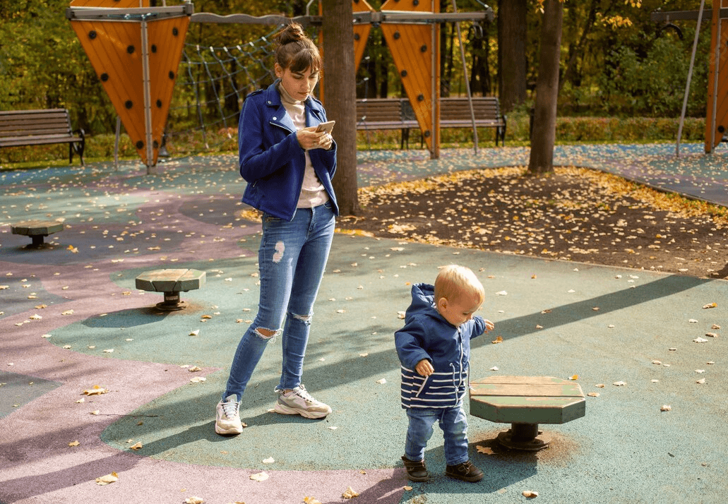 Az anya a mobilját nézi miközben gyermeke a játszótéren sétál.
